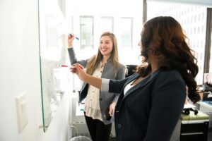 a woman standing at a whiteboard showing another woman some information. Both are dressed professionally and look happy.
