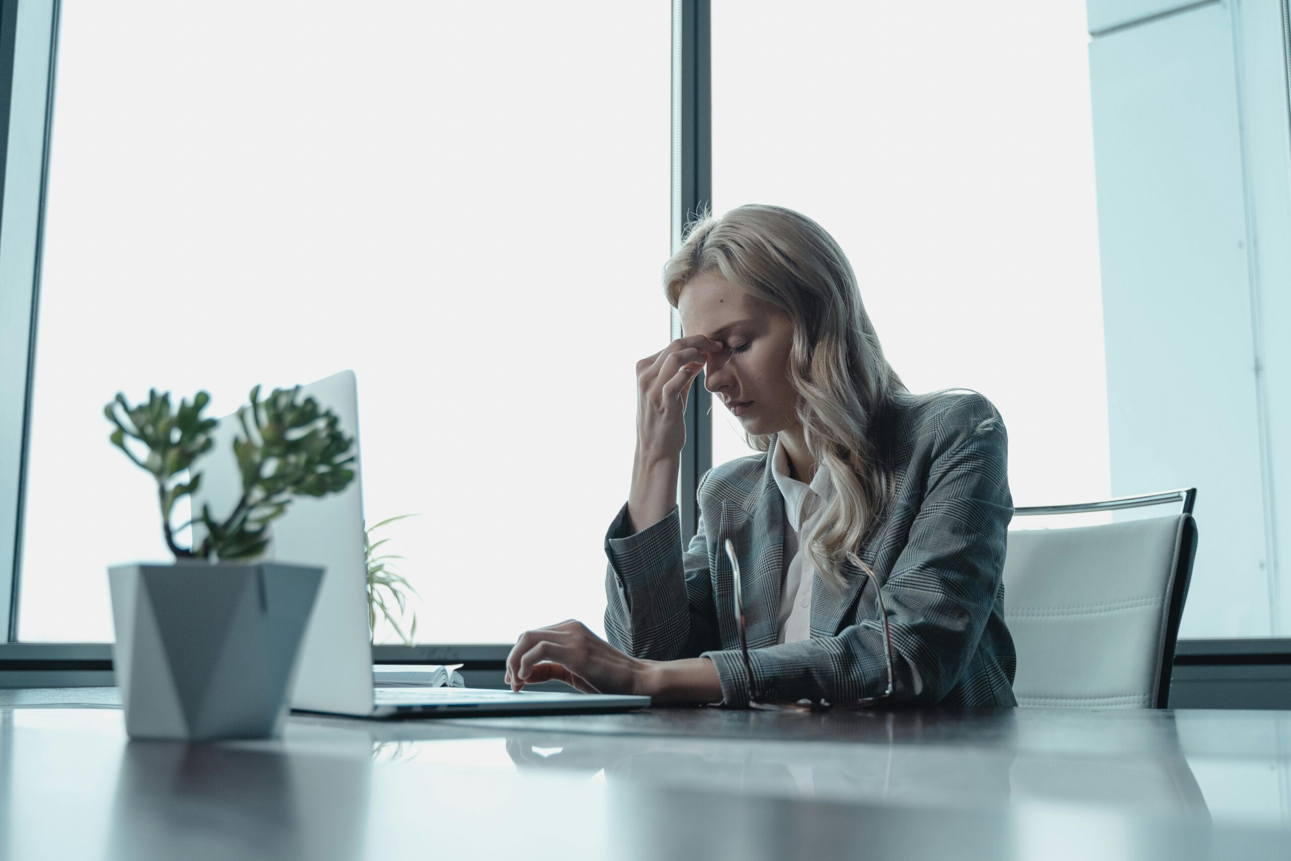 A woman in a grey suit sits at a desk with her head in her hands